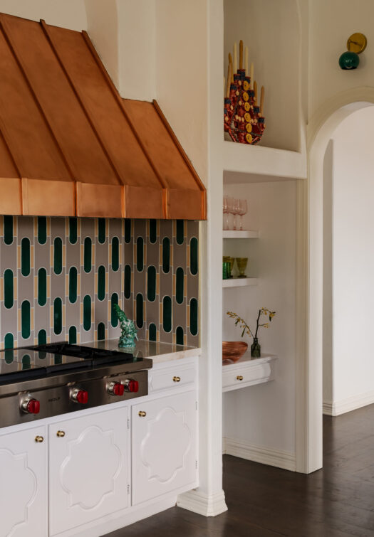 Cucina con cappa in rame, piastrelle verdi e bianche, mobili bianchi e una nicchia con mensole.